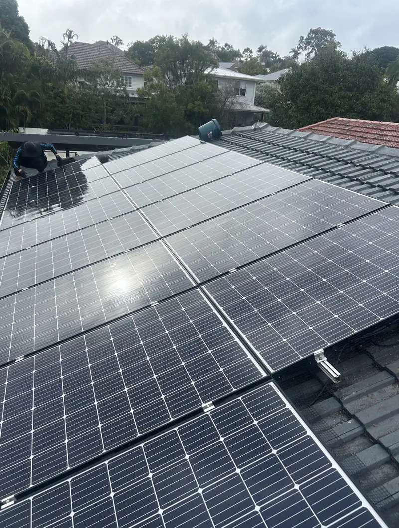 Solar panel bird proofing in Sunnybank: Technician crouched cleaning a large solar array on a grey tile roof under overcast sky