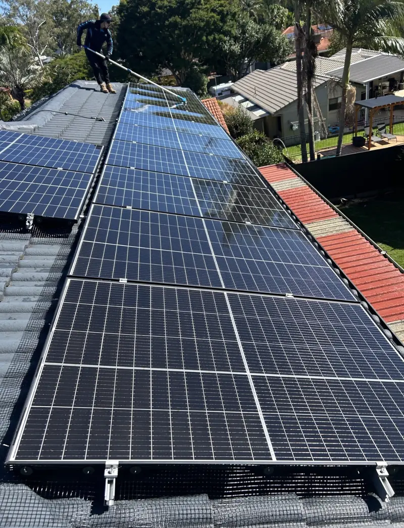 Solar panel bird proofing in The Gap: Worker cleaning long solar panel rows with a water-fed pole from a grey tile roof ridge