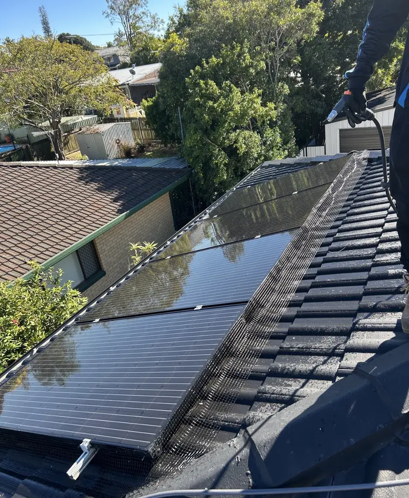 Solar panel bird proofing in Tingalpa: Installer aiming a spray gun over black solar panels edged with mesh on charcoal tiles